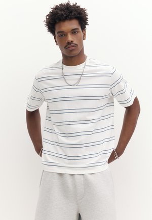 Junger Mann mit lockigem Haar, der ein weißes Kurzarmhemd mit dünnen horizontalen Streifen und hellgraue Hosen trägt, steht mit den Händen in den Taschen.