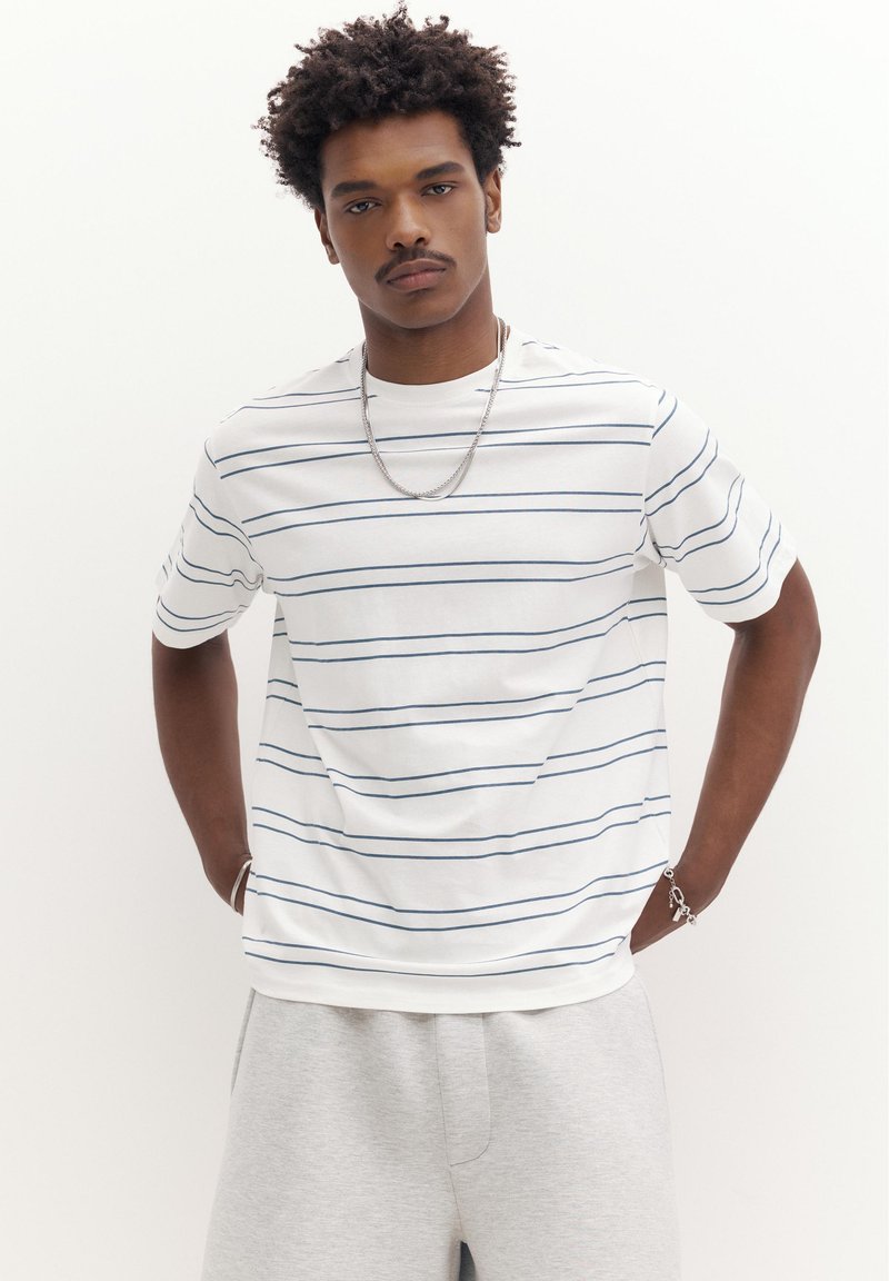 Jeune homme aux cheveux bouclés portant une chemise blanche à manches courtes avec de fines rayures horizontales et un pantalon gris clair, debout les mains dans les poches.