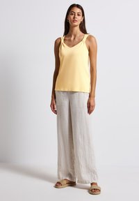 Gul ärmlös topp med V-ringning och knutdetaljer på axlarna, kombinerad med lösa beige linnByxor och vävda slidesandaler.