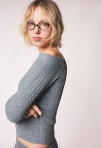 Sivkasta pletena pulover z odprtimi rameni, z rebrastimi detajli in pridobljenim dizajnom. Tekstura deluje mehka in prijetna, kar poudarja prijetno barvo.