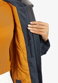 Personne tenant un manteau bleu marine ouvert montrant une doublure intérieure matelassée orange et une poche intérieure zippée.