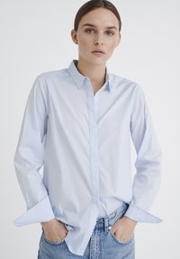Light blue long-sleeve shirt in cotton blend, with a button-down collar and folded cuffs, paired with blue jeans, displayed against a white background.