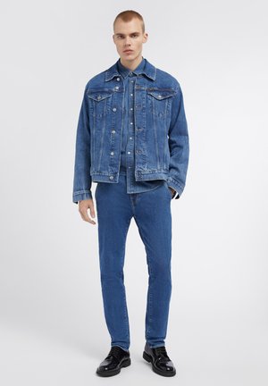 Giacca di denim con design classico, colore blu medio, chiusura con bottoni, tasche sul petto, abbinata a jeans blu coordinati e scarpe nere.