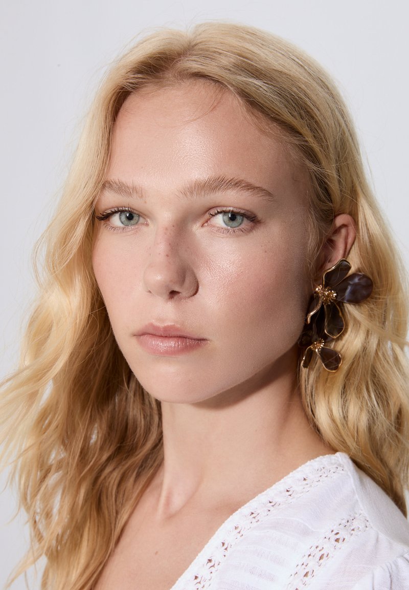 Blommiga örhängen i guldfärg med bruna genomskinliga kronblad och pärldetaljer, som bärs av en modell med långt blont hår och en vit texturerad topp.
