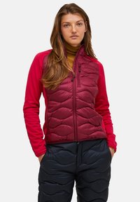 Jonge vrouw met lang haar, gekleed in een rode gesteppte jas, mosterdkleurige coltrui en zwarte gesteppte broek, met haar handen in haar zakken.
