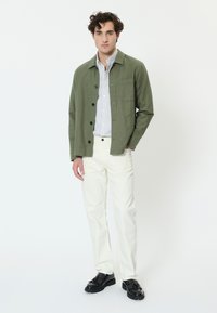Muž stojaci proti bielemu pozadiu, oblečený v olivovo zelenej zapínanej bunde, bielej košeli s pásikmi, bielych nohaviciach a čiernych kožených mokasínach.