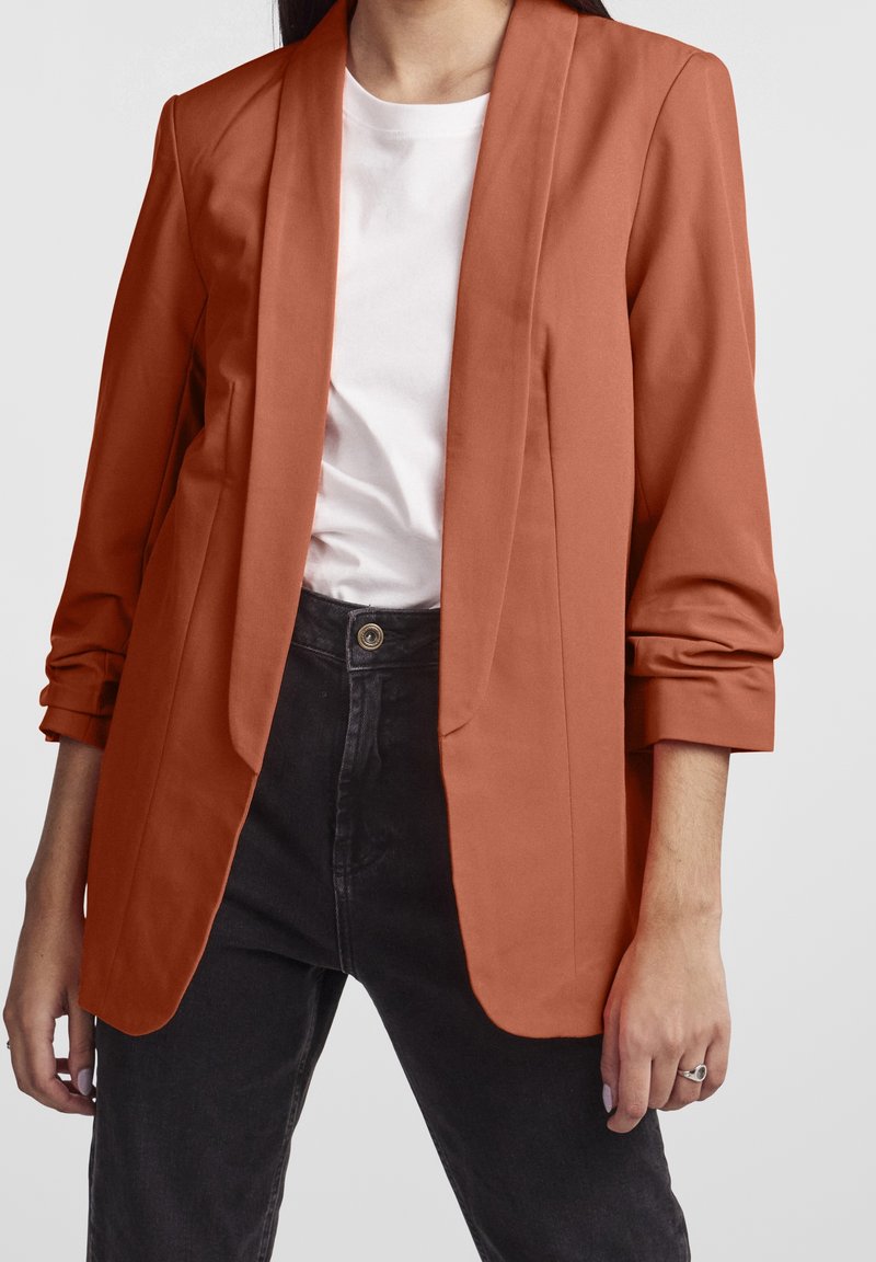 Femme portant un blazer couleur rouille sur une chemise blanche et un jean noir taille haute, avec les manches du blazer retroussées et une bague à la main gauche.