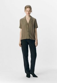 Camisa de manga corta verde olivo con cuello notch y botones en el frente, combinada con unos jeans negros de cintura alta y zapatos negros de punta.