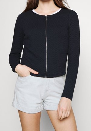Veste matelassée zippée marine à manches longues et col rond, associée à un short en denim clair. Présente un tissu texturé et une fermeture éclair argentée.