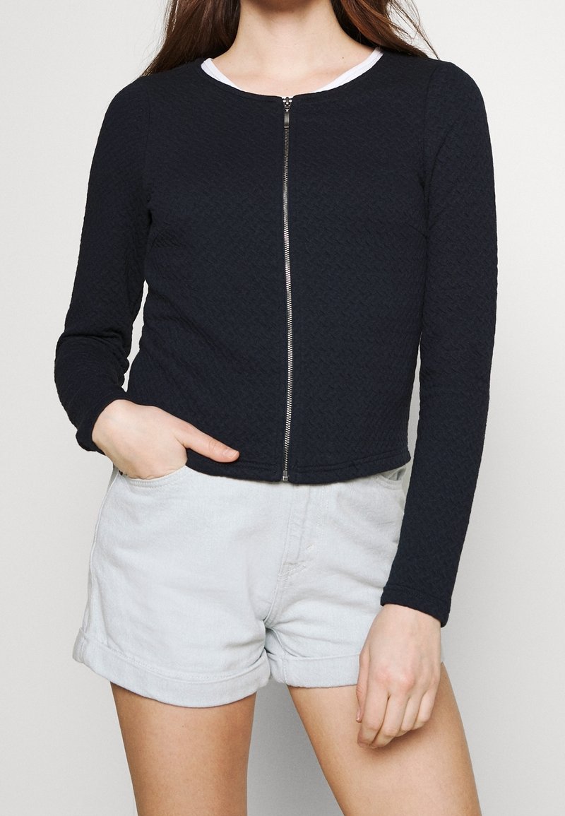 Navy quilted zip-up jacket with long sleeves and a round neckline, paired with light denim shorts. Features textured fabric and a silver zipper.