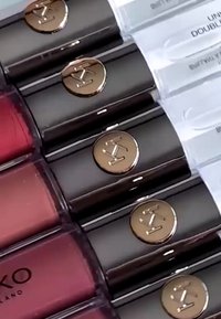 File di tubetti di rossetto di colore scuro con loghi dorati accanto a campioni rettangolari di diverse tonalità di balsamo per labbra etichettati per confronto.