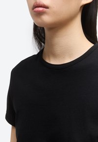 Κοντινό πλάνο ενός ατόμου που φοράει μια απλή μαύρη μπλούζα με στρογγυλή λαιμόκοψη, δείχνοντας τον λαιμό, το κάτω μέρος του προσώπου και το πάνω μέρος του στήθους σε φόντο λευκού.