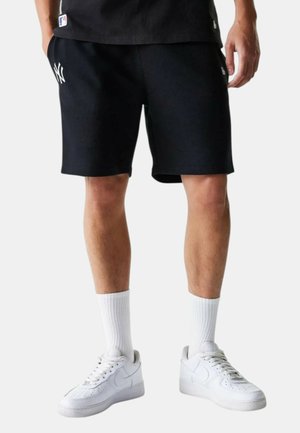 Hombre con pantalones cortos negros de los New York Yankees, calcetas blancas hasta la pantorrilla y zapatillas blancas Nike, de pie frente a un fondo claro y liso.