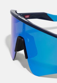 Blauw getinte zonnebril met een gestroomlijnd ontwerp, voorzien van een gebogen lens, zwarte hardware en getextureerde accenten op het montuur.