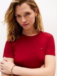 Camiseta roja de manga corta hecha de algodón, con un pequeño logo en el pecho. La modelo tiene el cabello ondulado y aretes de aro dorados.