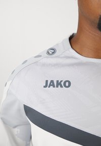 Grijze sportshirt van textuurstof, met een zwarte streep, een rond logo op de schouder en "JAKO" in het grijs gedrukt.