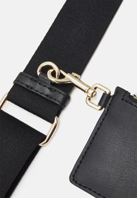 Cinghia in nylon nera con una chiusura in metallo dorato, dettagli in pelle nera e una superficie testurizzata. Presenta un attacco rettangolare per identificazione o decorazione.
