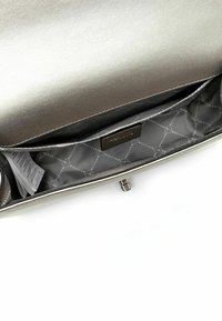 Interior de bolso metálico plateado con una textura suave, que cuenta con un compartimento principal y un pequeño bolsillo con una etiqueta de marca.