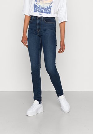 Personne portant un jean skinny bleu foncé, un t-shirt blanc avec un motif graphique et des baskets compensées blanches, debout devant un fond clair uni.