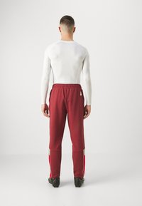 Rote Sporthose mit einem elastischen Bund, ausgestattet mit grauen und roten Seitenakzenten, kombiniert mit einem weißen langärmligen, enganliegenden Oberteil.