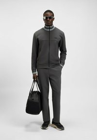 Grå atletisk träningsoverall med zip-up jacka som har ribbad krage, kontrasterande band och matchande byxor. Håller en svart duffelväska. Svarta sneakers.