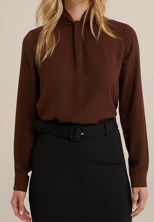 Kvinde iført en brun bluse med lange ærmer, stoppet ned i en sort høj-taljet nederdel med et sort bælte med en rektangulær spænde.