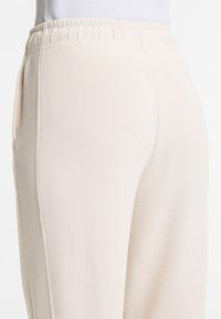 Light beige joggers with an elastic waistband, side pockets, and a smooth, soft texture. Features a fitted design with tapered legs.