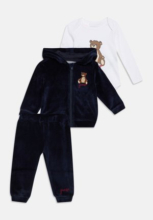 Ensemble bébé avec veste à capuche bleu marine et pantalon avec des patchs nounours, assorti à un body blanc à manches longues également décoré d’un nounours.
