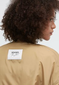 Beige Bomberjacke mit glatter Textur, ausgestattet mit einem weißen Stofflabel auf der Rückseite mit schwarzem Text. Lockige Haare sind seitlich sichtbar.