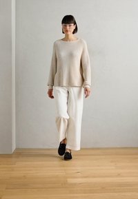 Beige neulepaita, jossa on ribbitekstuuri, yhdistettynä löysiin valkoisiin housuihin. Mallilla on päällään mustat kengät ja läpinäkyvät kehykset, ja hän seisoo puulattialla.