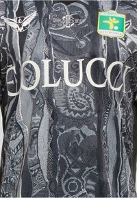 Dunkel gemustertes Sporttrikot mit weißem "GOLUCCI"-Text, einem Vogel-Logo auf der linken Seite und einem grün-gelben "Fussballclub Kitzbühel"-Patch auf der rechten Seite.