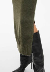 Robe en tricot vert olive avec un ourlet côtelé et des fentes latérales, associée à de longues bottes à talons en cuir noir au design épuré.