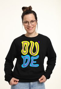 Felpa nera con testo audace e colorato che dice "DUDE" in giallo, viola e blu. Il materiale sembra morbido; il design è casual e rilassato.