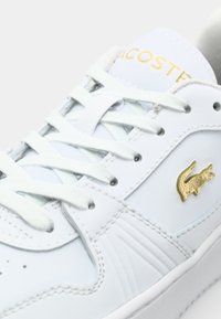 Balti odiniai kedai Lacoste su auksiniu logotipu, plokščiomis baltomis raištelėmis, tekstūruotais šonų akcentais ir lygiu paviršiumi.