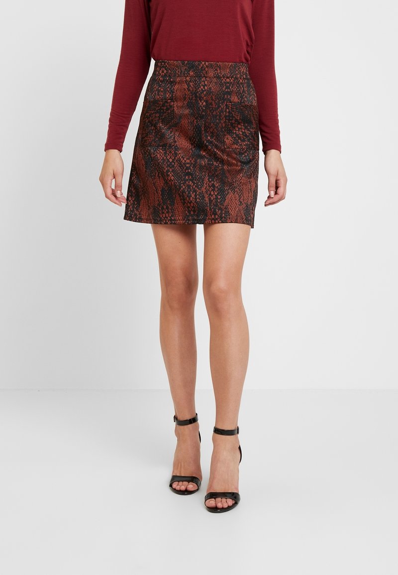 Femme portant une mini-jupe imprimé serpent rouge, un haut manches longues bordeaux et des sandales à talons hauts avec bride à la cheville noires, debout devant un fond blanc.
