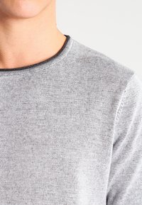 Světle šedý svetr, klasický výstřih, pletená textura, s jemnými variacemi v tloušťce nitě a žebrovaným výstřihem. Žádné výrazné vzory ani akcenty.