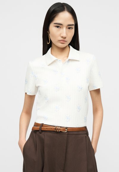 Jeune femme aux longs cheveux foncés portant une chemise blanche à manches courtes avec des motifs floraux bleu clair, un pantalon marron et une ceinture beige.