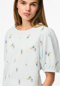 Top azzurro chiaro con trama, maniche corte a sbuffo, decorato con ricami floreali gialli e viola sparsi. Collo rotondo.