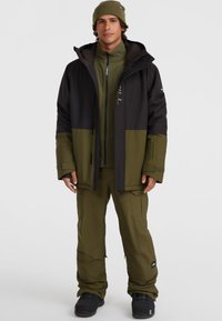 Veste imperméable verte et noire avec fermeture éclair, poignets ajustables ; assortie à un pantalon vert et des bottes noires. Modèle portant un bonnet.