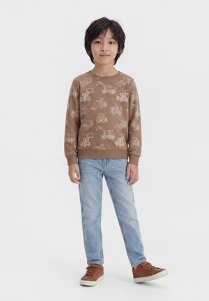 Dreng med mørkt hår iført en brun sweatshirt med print af byggepladskøretøjer, lyseblå jeans og brune sneakers, stående mod en hvid baggrund.