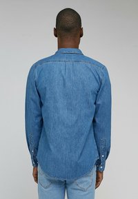 Man in een blauw spijkerhemd met lange mouwen en lichtblauwe jeans, staande met zijn rug naar de camera tegen een effen grijze achtergrond.