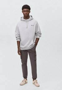 Un sweat à capuche gris clair avec une poche frontale et un logo en étoile. Associé à un pantalon cargo gris foncé et des baskets beiges. Le tissu semble doux et décontracté.