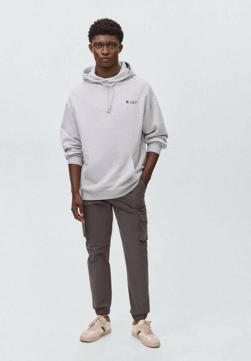 Un sweat à capuche gris clair avec une poche frontale et un logo en étoile. Associé à un pantalon cargo gris foncé et des baskets beiges. Le tissu semble doux et décontracté.