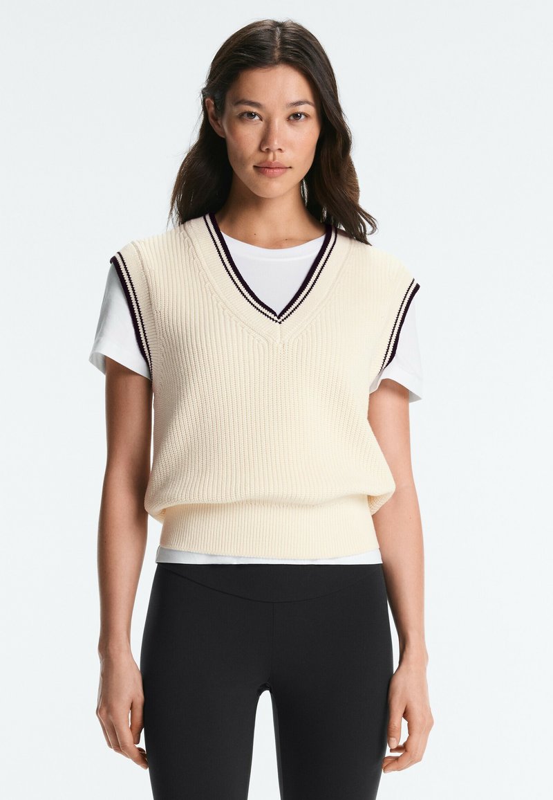Jeune femme aux longs cheveux foncés portant un gilet sans manches en maille crème sur un t-shirt blanc et un pantalon noir, debout devant un fond blanc.