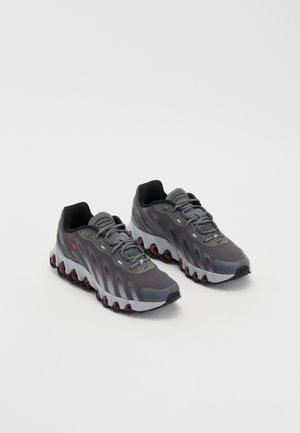 Paire de chaussures de sport gris et noir avec des accents rouges et des semelles amortissantes uniques sur un fond blanc uni, inclinées vers la droite.