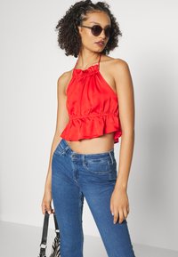 Top cropped vermelho com decote halter e bainha em camadas, combinado com jeans azuis ajustados. Modelo segura uma bolsa com riscas brancas e pretas, usando óculos de sol.