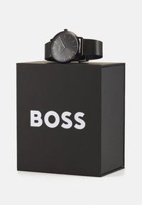 Musta metallkell minimalistliku ümmarguse näo javõrgust rihmaga, asetatud mustale karbile, millel on valge "BOSS" logo.
