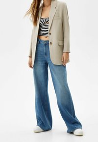 Blazer beige su top a righe bianche e nere, abbinato a jeans blu a vita alta e gamba larga, e sneakers bianche.