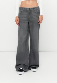 Wide-leg grey denim jeans with a faded finish, featuring five pockets, a button closure, and straight hems. Paired with black sneakers.