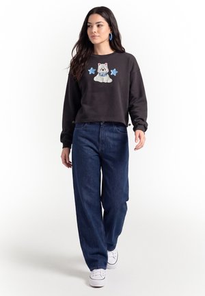 Kvinde går iført en sort sweatshirt med et design af en kat og stjerner, løse mørkeblå jeans, hvide sneakers og blå øreringe i form af ringe.